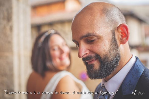 Foto 1026 de r2clic.com - Proyecto 365VecesTeQuiero (Fotos de boda y frases de amor) - Reportaje de boda en Garganta la Olla. Novio en primer plano y novia al fondo mirándole y sonriendo. Frase acompañando a foto: “Si como te amo a ti se amaran los demás… el mundo sería un lugar mejor” (Reportaje de boda en Garganta la Olla)